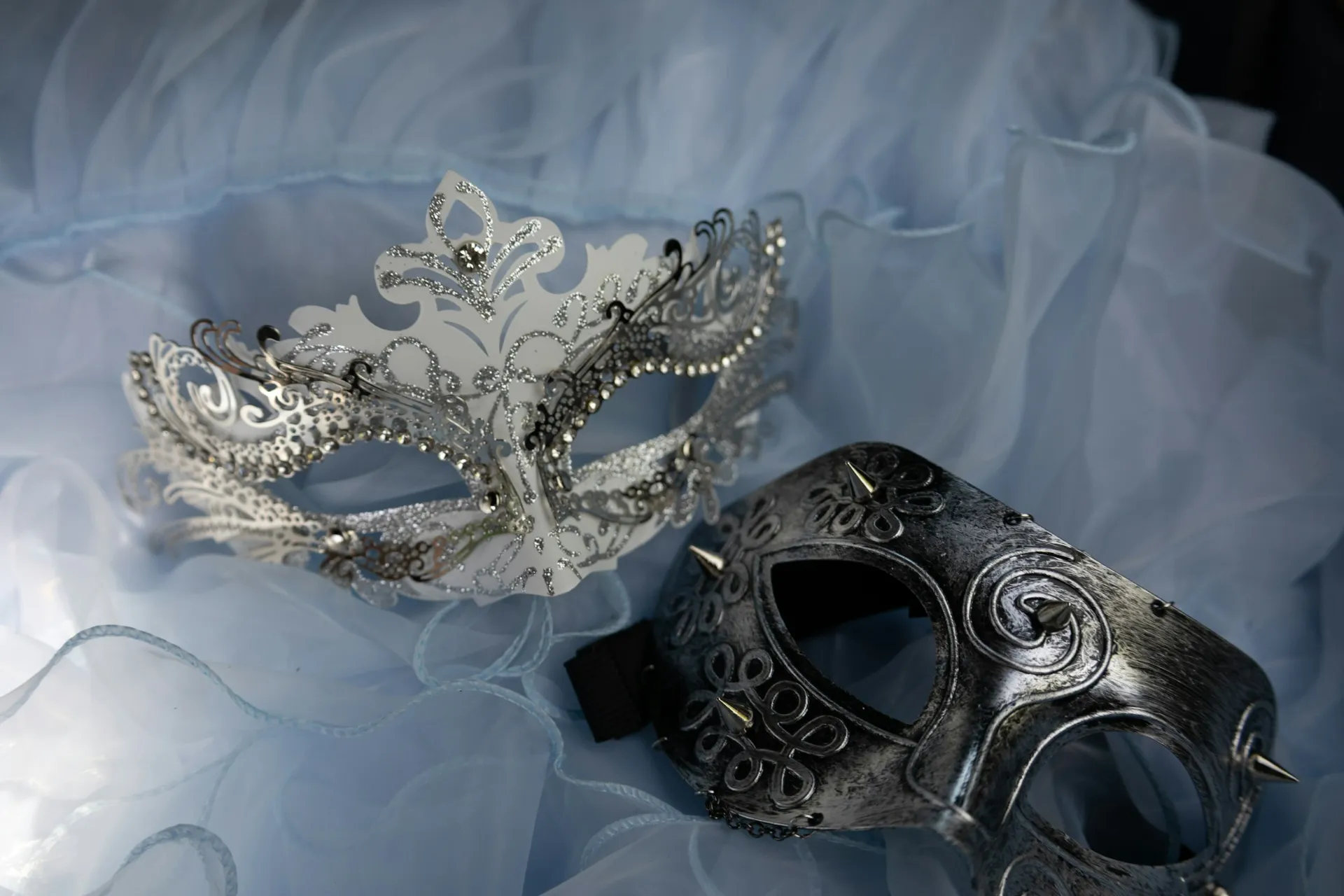 Two ornate masquerade masks, one silver and one dark, on pale blue fabric